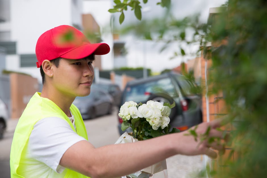 A delivery person wearing a red cap and a high-visibility yellow vest is seen reaching out to a residential brick wall to hand over a bouquet of fresh, white flowers, including roses and hydrangeas, a