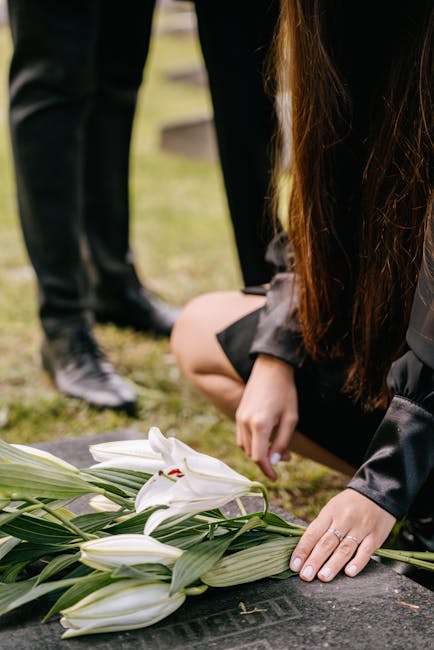 A close-up of a woman kneeling on the grass during a funeral, arranging a bouquet of white lilies with long green leaves on a dark stone memorial. The lilies are fresh, with smooth, white petals and p