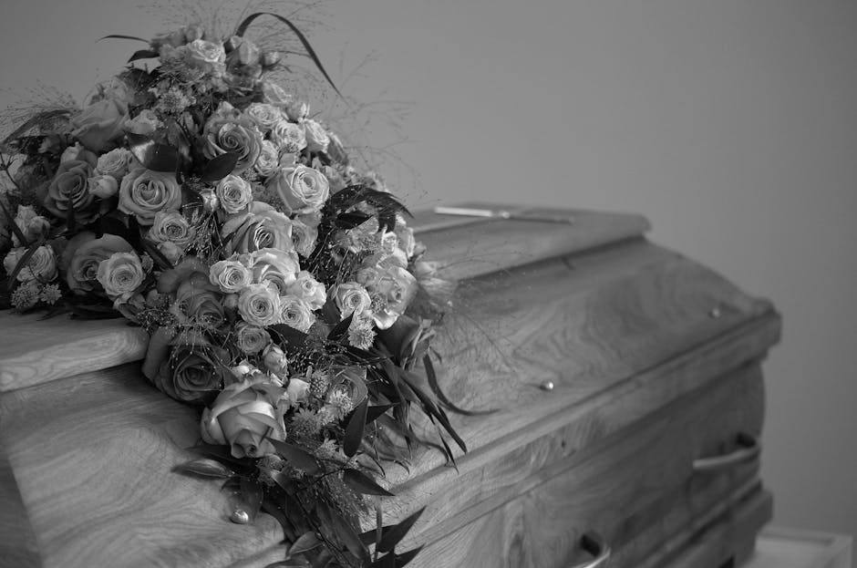 A large, elegant floral arrangement of freshly cut roses in various stages of bloom, with some fully open and others still budding, laid across the top of a closed wooden coffin. The bouquet includes 