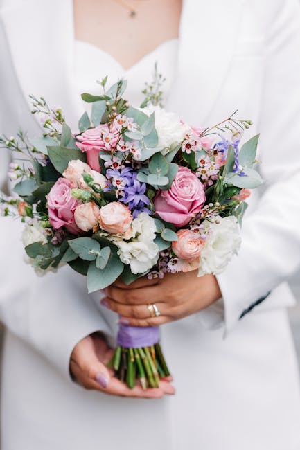 A person wearing a white outfit holds a bridal bouquet featuring a delicate arrangement of pink roses, lavender, white and cream flowers, complemented by sprigs of eucalyptus and small additional blos